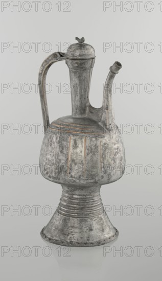 Jug aftabe, Islamic art; jug aftabe for water, 19th century, North Caucasus; vessel; height 30.0 cm, length 14.8 cm, diameter 12.0 cm, A metal vessel with a tall, rounded body and a flared base stands against a pale gray background; the vessel has a long curved handle and a narrow curved spout, a domed lid with a small decorative finial, and a textured surface showing areas of gray, silver, and traces of brownish tones, with worn and weathered patches and subtle vertical and horizontal ridges.