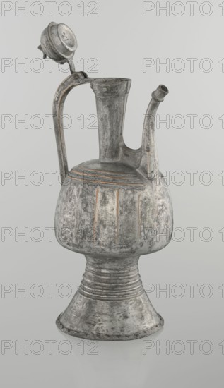 Jug aftabe, unknown provenance; Islamic art; jug used for water aftabe; 19th century; North Caucasus; vessel; height 30.0 cm, length 14.8 cm, diameter 12.0 cm, A metal vessel with a tall narrow neck, a curved spout, and a looped handle topped by a hinged lid, all showing a worn, textured surface. The body of the vessel is bulbous above a flared pedestal base with horizontal ridges where the body meets the stem. Vertical incised grooves decorate the rounded midsection, and concentric incised bands appear near the shoulder. The hinged lid sits open and is circular with a domed top. The spout is slightly curved and tapers toward its opening. The surface displays a mottled, aged appearance with visible areas of abrasion and patina. The visible colors are shades of gray, silver, off-white, and touches of muted brown or rust. The background is a smooth, light gray.