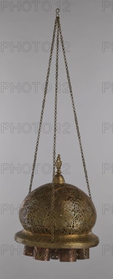 Lamp, 19th–20th century, Damascus, Syria. Metal oil lamp with cover decorated with a star motif; disc base with seven holes for glass sleeves glass oil containers missing. Arabic inscriptions; geometric ornament. Height 40 cm, diameter 32 cm, weight 2.4 kg. Culture: Islamic, Arabic., A hanging ornate metal lantern suspended by a few long chains from a single hook, with a domed top and a decorative finial; the lantern is predominantly brass-colored with darker bronze accents and features intricate openwork patterns and geometric filigree across the dome and lower rim, and a row of cylindrical decorative elements hanging beneath the rim.