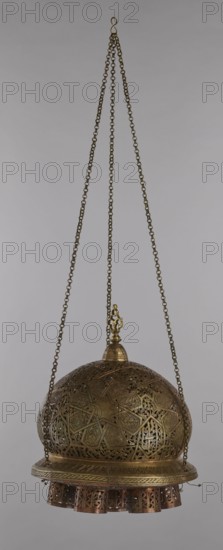Lamp, Islamic art, Arabic culture, metals, lighting, geometric ornament, Arabic inscriptions. Cover decorated with a star motif. Disc base with seven holes for glass sleeves glass oil containers missing. 19th–20th century, Damascus, Syria. Lamp; height 40 cm, diameter 32 cm, weight 2.4 kg., A hanging metal lantern suspended by a group of chains with an S-shaped hook at the top, the lantern has a domed, filigreed body with intricate openwork geometric and floral patterns, a small ornate finial at the dome peak, a decorative band around the lower edge, and a fringe of cylindrical cutout metal pieces at the bottom; visible colors include brassy gold, bronze, copper tones, and the neutral pale gray background.
