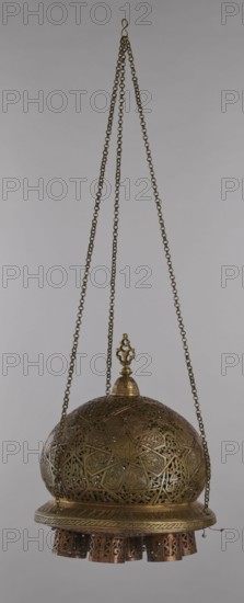Lamp, Islamic art, Arabic culture; metal oil lamp with Arabic inscriptions and geometric ornament. Cover decorated with a star motif. Disc base with seven holes for glass sleeves glass oil containers missing. 19th–20th century, Damascus, Syria. Height 40 cm; diameter 32 cm; weight 2.4 kg., A hanging metal lantern suspended by a group of chains, with a hooked top and three long linked chains converging at the hook, the chains and metal are brass-colored and aged, the lantern has a domed, perforated upper section with intricate geometric and floral openwork in brass tones, a decorative finial atop the dome with brass coloring, a circular rim below the dome with engraved patterns in a similar brass hue, and a skirt of several cylindrical, pierced pendants at the bottom that show darker copper and bronze tones; the background is a neutral light gray.