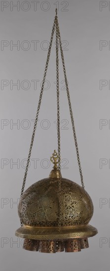 Lamp, Islamic art, Arabic, metal lighting fixture with geometric ornament and Arabic inscriptions. Cover decorated with a star motif. Disc base with seven holes for glass sleeves glass oil containers missing. 19th–20th century, Damascus, Syria. Lamp; height 40 cm, diameter 32 cm, weight 2.4 kg., A suspended decorative metal lantern with a domed, intricately pierced body hangs from three linked chains converging at a single hook; the chains and lantern are predominantly brass-colored with darker aged patina. The dome surface shows ornate geometric and curvilinear cutwork arranged in repeating patterns, with visible areas of sheen and shadow creating contrast between brighter golden highlights and deeper brownish tones. The top has a small finial with decorative openwork in the same brass color, and a patterned band encircles the base of the dome. Below the band, a ring of cylindrical perforated pendants hangs, showing a mix of brass and warmer reddish-brown metal hues, and the overall b...