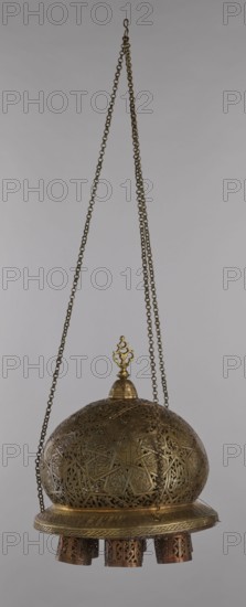 Lamp, Islamic art, Arabic culture, metals, lighting, light, geometric ornament, Arabic inscriptions, star ornament. Cover decorated with a star motif. Disc base with seven holes for glass sleeves glass oil containers missing. 19th/20th century, Damascus, Syria. Lamp; height 40 cm, diameter 32 cm, weight 2.4 kg., A hanging ornate metal object suspended by a few long linked chains against a plain light gray background, with a domed upper section covered in intricate filigree and geometric cutwork in brass and bronze tones, topped by a small decorative finial in a brighter golden hue; a circular rim in similar metallic tones below the dome features engraved patterns and supports a few short cylindrical dangling elements with pierced designs showing darker coppery and brownish interiors.