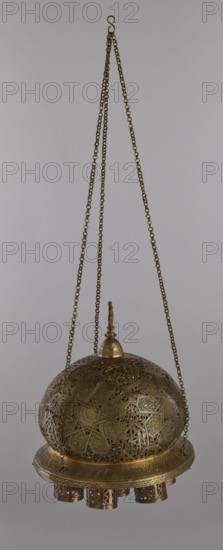 Lamp, Islamic art, Arabic culture, metals, lighting, geometric ornament, Arabic inscriptions. Cover decorated with a star motif. Disc base with seven holes for glass sleeves glass oil containers missing. 19th–20th century, Damascus, Syria. Lamp; height 40 cm, diameter 32 cm, weight 2.4 kg., A hanging decorative metal lamp suspended by a few long linked chains with an S-shaped hook at the top; the lamp has a domed, perforated body with intricate geometric and floral cutwork patterns and a small finial at its peak; a circular band forms the lower edge of the dome and beneath it hang a group of short cylindrical metal fixtures with openwork designs; visible colors include brassy gold tones on the metal and darker bronze or brown accents against a neutral light gray background.