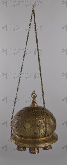 Lamp, Islamic art, Arabic culture; metal lighting fixture with geometric ornament and Arabic inscriptions. Cover decorated with a star motif. Disc base with seven holes for glass sleeves glass oil containers missing. 19th–20th century, Damascus, Syria. Lamp; height 40 cm, diameter 32 cm, weight 2.4 kg., A hanging metal lantern suspended by a long decorative chain, with a hook at the top and a pendant finial above a domed, perforated body; the lantern features intricate geometric and floral openwork patterns across the dome and band, and a circular base with short cylindrical feet that also have cutout designs. The visible colors are shades of brass and bronze, with warm golden brown metallic tones and subtle darker patina in the recessed areas, set against a plain light gray background.