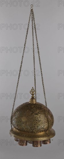 Lamp, Islamic art, Arabic culture; metal lamp with Arabic inscriptions and geometric ornament including a star motif on the cover. Disc base with seven holes for glass sleeves glass oil containers missing. 19th–20th century, Damascus, Syria. Height 40 cm, diameter 32 cm, weight 2.4 kg., A hanging metal lantern suspended by a group of chains, with a hooked loop at the top and three chains descending to a decorative finial; the lantern body is a domed, perforated and intricately patterned structure with geometric and floral openwork, showing metallic tones of brass and bronze with areas of darker patina and hints of copper; a circular rim supports a few short cylindrical pendants with pierced patterns beneath the dome; the background is a plain neutral gray.