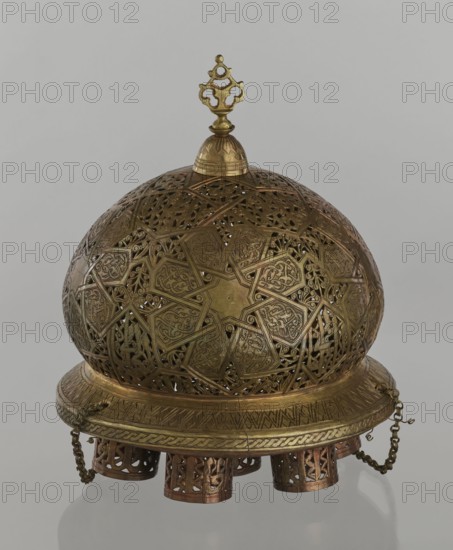 Lamp, Islamic art, Arabic culture; metal lighting vessel with geometric ornament and Arabic inscriptions. Cover decorated with a star motif. Disc base with seven holes for glass sleeves glass oil containers missing. 19th/20th century, Damascus, Syria. Lamp; height 40 cm, diameter 32 cm, weight 2.4 kg., A decorative domed metal object with intricate openwork patterns and filigree, showing a group of interlocking geometric and curvilinear designs across its surface in metallic gold and brass tones; a small ornate finial tops the dome in matching gold, and the dome sits on a circular base with engraved decorative bands and braided detailing in gold and brass; hanging chains and a few cylindrical perforated attachments in darker copper and bronze tones are visible beneath the base, and the object rests against a plain light gray background.