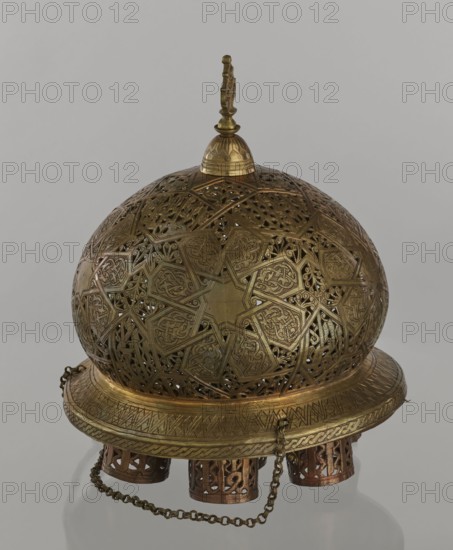 Lamp, Islamic art, Arabic culture; metal oil lamp with Arabic inscriptions and geometric ornament. Cover decorated with a star motif. Disc base with seven holes for glass sleeves glass oil containers missing. 19th–20th century, Damascus, Syria. Height 40 cm, diameter 32 cm, weight 2.4 kg., A rounded ornate metal object with a domed top and a finial, showing an intricate pierced lattice pattern across its surface and geometric star motifs, predominantly metallic gold and brass in color with some darker bronze tones; a wide circular base with engraved decorative bands supports the dome, and a short decorative chain of matching metallic color hangs from the base and loops around, while a few cylindrical openwork supports in a darker bronze sit beneath the base against a neutral light gray background.