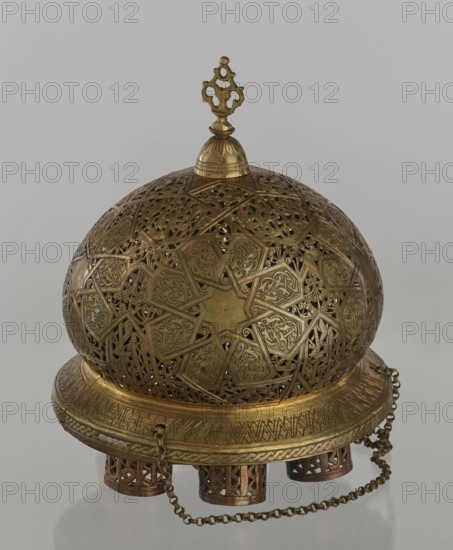 Lamp, Islamic art, Arabic culture; metal oil lamp with Arabic inscriptions and geometric ornament; cover decorated with a star motif; disc base with seven holes for glass sleeves glass oil containers missing; 19th–20th century, Damascus, Syria; height 40 cm, diameter 32 cm, weight 2.4 kg., A domed metal object with intricate openwork and ornate geometric and curvilinear patterns covering its surface, topped by a decorative finial; the piece sits on a wide circular base with engraved banding and a braided border, supported by a group of short cylindrical feet with cut-out designs, and a metal chain draped around the base. The visible colors are gold and brass tones against a neutral gray background.