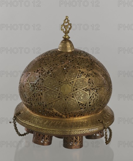 Lamp, creator unknown, Islamic art, Arabic culture; metal oil lamp with geometric ornament and Arabic inscriptions; cover decorated with a star motif; disc base with seven holes for glass sleeves glass oil containers missing. 19th–20th century, Damascus, Syria. Height 40 cm; diameter 32 cm; weight 2.4 kg., A rounded, ornate metal object with a domed lattice upper section and a circular base, primarily gold and brass in color with darker bronze accents; the dome features intricate geometric and floral cutwork with a star-shaped motif and repeating patterned panels, and a decorative finial sits atop a small rounded cap; the base has engraved banding and a braided decorative rim with a few hanging chain pieces, and the whole piece rests on a group of short cylindrical feet with pierced patterns, all set against a neutral light gray background.