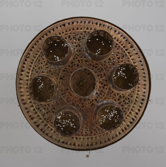 Lamp, 19th–20th century, Damascus, Syria. Metal Islamic lamp with Arabic inscriptions and geometric ornament; cover decorated with a star motif. Disc base with seven holes for glass sleeves glass oil containers missing. Height 40 cm, diameter 32 cm, weight 2.4 kg., A round, ornate object viewed from above with a predominantly brown and bronze color palette, featuring an outer rim and a patterned interior with geometric lattice work and carved scroll-like motifs; a central circular area is surrounded by a group of several hollow cylindrical cups or compartments with perforated sides that reveal darker brown interiors; the surface shows variations in brown tones with hints of darker patina and slight metallic sheen, and small metal hooks or loops extend from the rim at a few points against a neutral light gray background.