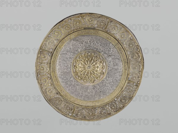 Mirror frame, 19th century, possibly Turkey or Iran. Metal; weight 685 g, diameter 25.5 cm, height 1.2 cm. Islamic art mirror frame with plant motifs and geometric ornament, featuring a 6-pointed star Solomon's Seal and stylized plant patterns arranged geometrically., A circular ornate metal plate with intricate filigree and embossed floral patterns, showing a central rosette with layered petal-like openwork, surrounded by concentric bands of detailed vine and leaf motifs; the plate displays a mix of metallic gold and silver tones, with areas of darker shadowing and light reflecting off the raised decorations, set against a neutral pale gray background.