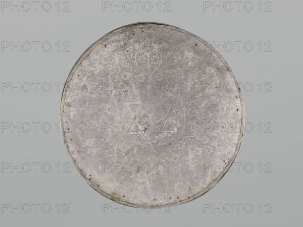 Mirror frame, scrap - purchase provenance; Islamic art; mirror frame with plant motifs and geometric ornament including 6-pointed star Solomon's Seal; stylized plant patterns in a geometric arrangement; 19th century; possibly Turkey or Iran; weight 685 g; diameter 25.5 cm; height 1.2 cm., A large round metal object fills the frame, shown against a solid light gray background. The object has a metallic silvery surface with an ornate engraved pattern covering its face, featuring swirling vines and floral motifs arranged symmetrically. A subtle six-pointed geometric design is visible near the center, integrated into the engraved decoration. The rim is slightly raised and shows small evenly spaced holes and minor darker spots and discolorations around its edge. The overall color palette is silvery gray with hints of darker gray in the engraved lines and some faint brownish tarnish near parts of the rim.