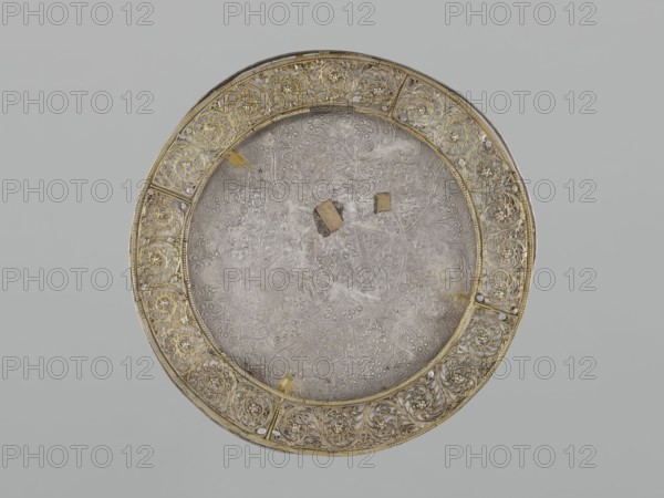 Mirror frame, maker, 19th century; possibly Turkey or Iran; Islamic art mirror frame with decoration of a 6-pointed star Solomon's Seal, stylized plant motifs in a geometric arrangement; geometric ornament, plant motifs, star iconography; materials unspecified; weight 685 g; diameter 25.5 cm; height 1.2 cm., A round decorative plate with an ornate border and a patterned central area sits against a pale gray background. The border features a repeating floral and scrollwork design in metallic gold and silver tones with small raised bead-like accents. The central area has an intricate embossed floral and geometric pattern in muted silver-gray with areas that appear slightly tarnished or dusty. A few small rectangular pieces in brownish-beige rest on the central surface. There are small spots of pale yellow discoloration on the central area and along the inner edge of the border. The overall color palette includes gold, silver, gray, beige, brown, and pale yellow.