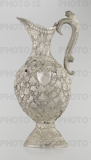 Jug, late 19th century, Northern India, Iran, silver vessel, height 43 cm, weight 1795 g. A tall silver jug with a pear-shaped base and a tall, conical body tapering upward into a wide spout. The entire surface is richly decorated with plant motifs—leaves, twigs, tendrils, carnations and other flowers. On one side of the belly is a blank shield. The handle is ornamented with images of spotted animals with large ears and open mouths; a hinge at the upper edge of the handle indicates the vessel originally had a lid. Zoomorphic decoration and coats of arms motifs present; categorized as Islamic art., A tall ornate metal pitcher with an intricately worked surface covered in floral and leaf patterns, featuring a curved handle with sculpted animal head details and decorative motif engravings, a flared spout with patterned rim, a rounded body with a shield-shaped smooth area, and a flared pedestal base with repeating ornamental borders; the visible colors are silvery metallic tones and subtle...