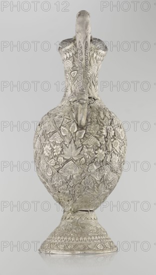 Jug, purchase, Islamic art, jugs, zoomorphism, coats of arms, floral motifs, plant ornament, leaves, carnations, plant tendrils. A tall silver jug with a pear-shaped base. The tall, conical base tapers upward, smoothly merging into a wide spout. The entire vessel is richly decorated with plant motifs—leaves, twigs, and various species of flowers. In the center of the belly wall, on one side only, is a blank shield. The jug's handle is decorated with images of spotted animals with large ears and open mouths. A hinge at the upper edge of the handle indicates the vessel originally had a lid. Late 19th century, possibly Northern India or Iran; height 43 cm, weight 1795 g., An ornate metal vessel with a long, curved neck and bulbous body sits upright on a flared base, all covered in dense raised floral and leaf patterns; the surface shows intricate carved motifs, textured bands, and a sculpted spout that projects from the neck area, with a small decorative element beneath it; the piece appe...