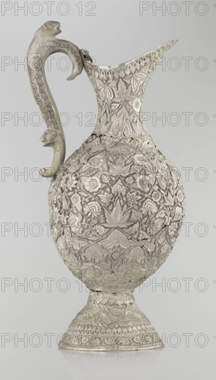 Jug, late 19th century, Northern India possibly / Iran possibly. Tall silver jug with a pear-shaped body and tall, conical neck tapering to a wide spout. Entire surface richly decorated with plant motifs—leaves, twigs, carnations and other flowers, and plant tendrils. On one side of the belly is a blank shield. Handle decorated with spotted animals with large ears and open mouths; hinge at upper edge of handle indicates the vessel originally had a lid. Height 43 cm; weight 1795 g. Styles/features: Islamic art; zoomorphism; coats of arms; floral ornament., A single ornate metal pitcher with an elongated spout and a curved handle sits against a plain light gray background; the pitcher is covered in dense raised floral and leaf patterns all over its bulbous body, neck, and flared base, and the handle features carved animal head motifs and repeating circular decorations. The surface appears metallic with tones of silver and muted gray, with subtle darker gray shadows in the recesses of the...