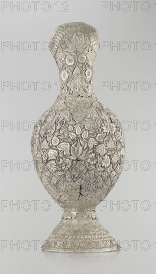 Jug, late 19th century, Northern India, or Iran, silver vessel, height 43 cm, weight 1795 g. A tall silver jug with a pear-shaped body and a tall conical neck that tapers upward into a wide spout. The entire surface is richly decorated with plant motifs—leaves, twigs, various flowers including carnations, and plant tendrils. On one side of the belly is a blank shield coat of arms. The handle is ornamented with zoomorphic figures: spotted animals with large ears and open mouths. A hinge at the upper edge of the handle indicates the vessel originally had a lid., A tall ornate vessel with a narrow neck and rounded body standing on a flared foot, entirely covered in intricate raised floral and leaf patterns, with visible seams and a few cracks in the surface; the surface appears metallic with shades of silver and pale gray, and the background is a smooth light gray.