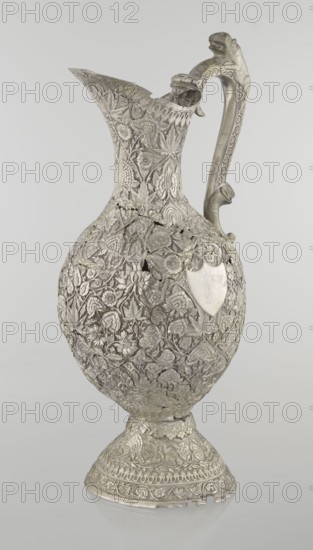 Jug, late 19th century, Northern India, / Iran, — silver, height 43 cm, weight 1795 g. A tall silver jug with a pear-shaped body and tall conical neck that tapers upward into a wide spout. The entire vessel is richly chased with plant motifs — leaves, tendrils, twigs, carnations and other flowers. On one side of the belly is a blank shield. The handle is ornamented with spotted animals with large ears and open mouths. A hinge at the upper edge of the handle indicates the vessel originally had a lid., A tall ornate metal pitcher stands against a plain light gray background; the pitcher is covered in dense raised floral and leaf patterns with textured surfaces and has a curved handle topped with an animal-like figure and a decorative spout area. The object appears in silver-gray tones with areas of darker gray shadowing and subtle highlights, and there is a smooth shield-shaped blank panel on one side that contrasts with the intricate decoration. Overall the visible colors are silver-gra...