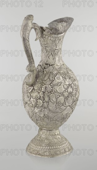 Jug, late 19th century, Northern India, or Iran, silver vessel, height 43 cm, weight 1795 g. A tall silver jug with a pear-shaped body and a tall, conical neck that tapers upward into a wide spout. The entire surface is richly chased with plant motifs—leaves, twigs, tendrils, carnations and other flowers. On one side of the belly is a blank shield. The handle is decorated with spotted animals with large ears and open mouths; a hinge at the upper edge of the handle indicates the jug originally had a lid. Style: Islamic art; motifs: zoomorphism, coats of arms, floral ornament., A single ornate metal pitcher with a curved handle and a flared spout is shown against a plain light gray background; the pitcher has an overall silvery-gray color with a slightly darker patina in recessed areas and is covered in dense raised floral and leaf patterns, including stylized blossoms, vines, and textured foliage across the body, neck, and base, while the handle features repeating decorative motifs and ...