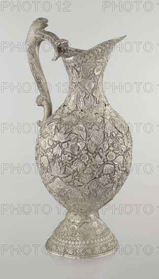 Jug, late 19th century, Northern India, Iran, silver vessel, height 43 cm, weight 1795 g. A tall silver jug with a pear-shaped body and tall conical neck that tapers upward into a wide spout. The entire vessel is richly decorated with plant motifs—leaves, twigs, carnations, and tendrils. On one side of the belly is a blank shield. The handle is decorated with images of spotted animals with large ears and open mouths; a hinge at the upper edge of the handle indicates the vessel originally had a lid. Style: Islamic art; themes: zoomorphism, coats of arms, floral ornament., The image shows an ornate metal pitcher with a high curved handle and a flared spout, covered all over in dense raised floral and foliage patterns and decorative motifs. The surface appears textured with carved leaves, flowers, and vine-like designs forming an overall repeating pattern that wraps around the rounded body and continues onto the pedestal base. The handle is sculpted with decorative elements and a small pr...