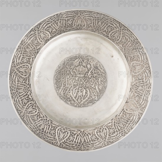 Plate, Habib Joo & Sons maker, floral and geometric ornament, crowns, medallion filled with floral ornament with an interwoven crown and the number XXXI., inscription 1868, India, Srinagar; vessel; height 2.5 cm, width 28.8 cm, weight 440 g., A round metallic plate with a decorative raised rim and a shallow central well, showing a central circular medallion of intricate carved vine and leaf patterns and a wide outer band of repeating interlaced floral and geometric motifs; the surface is primarily silver with areas of darker patina in the engraved recesses, and the background around the plate is a pale gray.