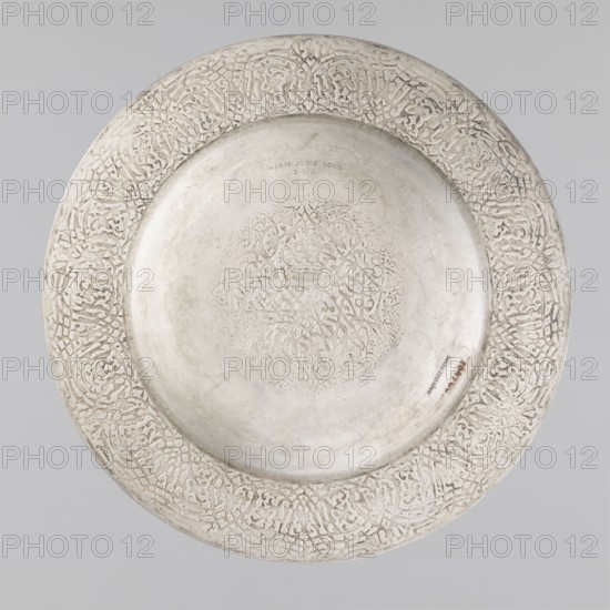 Plate, Habib Joo & Sons craftsman, floral and geometric ornament, crowns, medallion filled with floral ornament with an interwoven crown and the number: XXXI., 1868, India, Srinagar, vessel, height 2.5 cm, width 28.8 cm, weight 440 g, A round, shallow metal dish with an ornate raised border featuring an intricate repeating carved pattern, a smooth central well with a faintly incised circular motif, and an overall silvery-gray surface showing subtle variations in sheen and slight areas of darker patina.