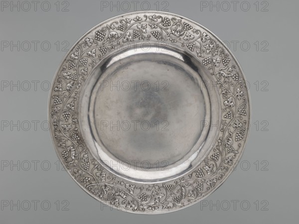 Platter, attributed to an, Qing dynasty 1644–1911, 19th–20th century, China. Ceramic decorative vessel depicting grapes, vines, and squirrels. Diameter 35.0 cm; weight 1170 g., A round metal plate with a reflective central basin surrounded by a wide decorative rim featuring an ornate vine pattern with leaves and clustered berry shapes; the plate appears in shades of silver and grey against a muted grey background.