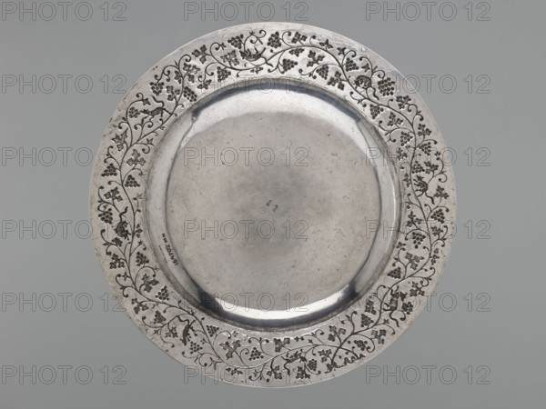 Platter, purchased provenance, Chinese art, Chinese culture, Qing 1644–1911, grapes, decorative vessel, squirrels, vines, 19th–20th century, China, vessel, weight 1170 g, diameter 35.0 cm, A round metallic plate with a broad decorative rim and a slightly recessed central well, showing a smooth reflective surface and visible scratches and wear; the rim features an ornate vine-and-grape pattern of engraved curling stems, leaves, and clusters arranged around the circumference; the metal appears silver-gray with darker gray accents in the engraved areas, and the background is a uniform muted gray.