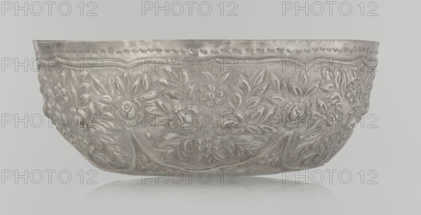 Bowl, Islamic art, Turkish, Ottoman dynasty 1299–1922, floral ornament, plant ornament, wall decorated with floral motifs in the valance, late 19th century, Turkey, vessel, weight 295 g, height 7.0 cm, diameter 18.0 cm, A shallow decorative bowl shown from the side with embossed floral and leaf patterns covering its exterior in a metallic silver-gray color, placed against a pale gray background with a faint reflective surface below; the bowl has a scalloped rim and textured surface with highlights and subtle darker tarnished areas.