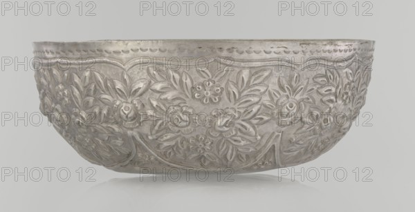 Bowl, late 19th century, Turkey, Ottoman dynasty 1299–1922; ceramic vessel decorated with floral and plant ornament—wall decorated with floral motifs in the valance; weight 295 g; height 7.0 cm; diameter 18.0 cm; Islamic art, Turkish., A shallow, rounded metal bowl with a reflective silver surface sits against a pale gray background; its exterior is embossed with an ornate floral relief featuring a group of stylized flowers and leaves arranged in repeating panels, with raised scalloped edging near the rim and textured areas between the raised motifs, showing a few variations in sheen and subtle darker patina in crevices.