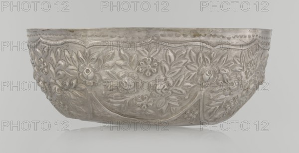 Bowl, late 19th century, Turkey; Turkish, Ottoman dynasty 1299–1922; ceramic vessel decorated with floral and plant motifs, wall-like valance decorated with floral motifs; height 7.0 cm, diameter 18.0 cm, weight 295 g., A shallow rounded metal bowl with ornate raised floral and leaf relief all around its exterior, showing a repeating scalloped border near the rim and decorative curved panels; the surface appears metallic silver with areas of slightly darker patina and subtle brownish spots, set against a light gray background with a faint reflection below.