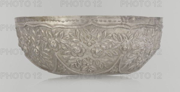 Bowl, late 19th century, Turkey; Ottoman dynasty 1299–1922; Islamic art, Turkish; vessel; wall decorated with floral motifs in the valance; floral and plant ornament; weight 295 g; height 7.0 cm; diameter 18.0 cm., A metallic bowl with embossed floral patterns covering its exterior, showing raised flowers and leaves in a repeating decorative design; the surface appears textured with hammered and sculpted details and a scalloped rim; visible colors include silver, gray, and hints of off-white, set against a pale gray background.