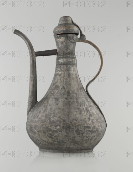 Jug aftabe, Islamic art, Islamic culture, jug for water, geometric ornament, 19th century, Caucasus, or Central Asia, vessel; height 31.5 cm, width 20.5 cm, diameter 18 cm, A metal pitcher with a rounded base, a narrow neck, and a hinged domed lid sits against a pale background; the surface shows a weathered, uneven texture with areas of dark gray, lighter gray, and muted brownish tones, and a curved handle with similar patina on one side and a long spout on the other, with visible wear and slight discoloration along edges.