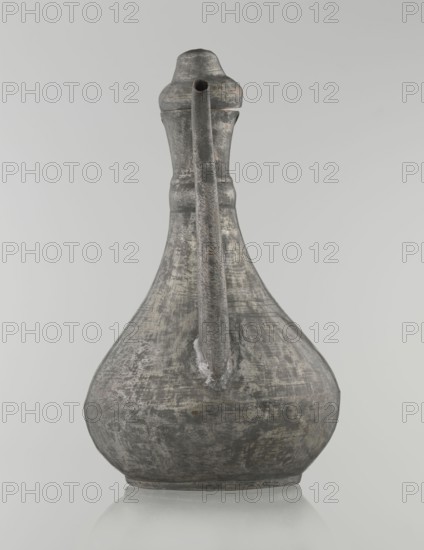Jug aftabe, repossession provenance; Islamic art; Islamic culture; jug for water; geometric ornament; 19th century; possibly Caucasus or Central Asia; vessel; height 31.5 cm, width 20.5 cm, diameter 18 cm, A single tall vessel with a rounded base and a narrow neck, shown from the front, with a long curved handle running from the upper neck down to the body and a short spout at the top; the surface shows a weathered, textured patina with visible abrasions and worn areas. The colors visible are shades of gray, dark gray, and hints of muted brown, with lighter gray highlights where the surface appears more worn. The background is a smooth pale gray and the vessel sits on a reflective pale surface that mirrors its lower shape.