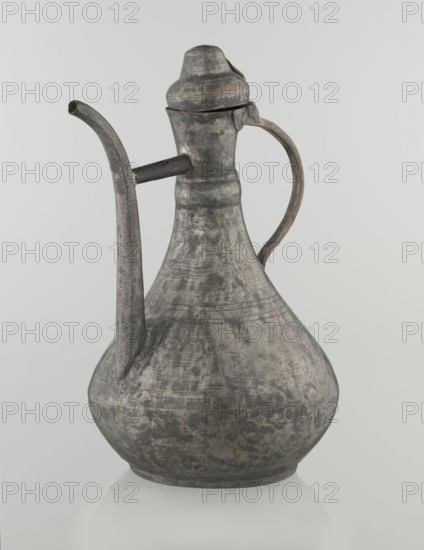 Jug aftabe, Islamic art, Islamic culture, jug for water, geometric ornament, 19th century, possibly Caucasus or Central Asia, vessel; height 31.5 cm, width 20.5 cm, diameter 18 cm, A single metal pitcher with a rounded base and a tapered neck sits against a pale background; the surface of the pitcher shows a weathered, mottled appearance in shades of gray with areas of darker gray and hints of brownish tones, and it has a curved handle attached to the neck and a long spout extending from the shoulder, plus a lid with a rounded top.