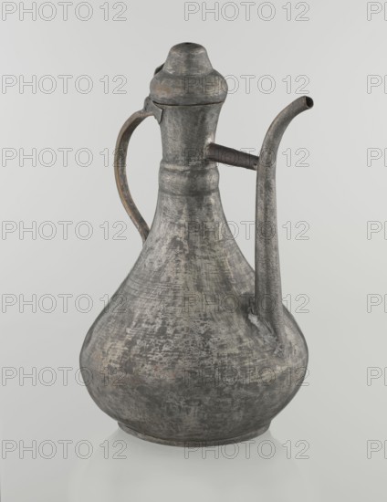 Jug aftabe, Islamic art, Islamic culture, jugs, water, geometric ornament, 19th century, Caucasus, Central Asia, vessel; height 31.5 cm, width 20.5 cm, diameter 18 cm, A metal pitcher with a bulbous base and a narrow neck sits against a pale background, showing a weathered surface with mottled shades of gray and patches of darker and lighter gray, along with small areas of muted brownish and rusty tones; it has a curved handle attached near the neck and a long curved spout extending from the shoulder, both sharing the same textured, aged gray appearance, and a fitted lid with a rounded top that matches the overall metallic, worn finish.