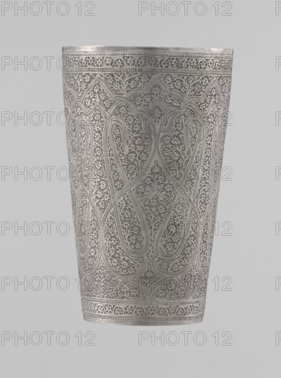 Mug, 19th century, North India, Mughal/Indian style. Silver mug densely decorated with horror vacui plant and floral motifs, featuring large symmetrically extending lines on the side wall forming shapes reminiscent of flower buds. Upper and lower rims decorated with horizontal ornamental lines. Interior gilded and polished. Weight 190 g, height 12.8 cm, diameter 7.8 cm., A tall, cylindrical metal vessel with intricate, repeating engraved patterns covering its surface, featuring interlacing curved motifs and dense floral or foliage designs, with decorative bands at the top and bottom; the visible colors are shades of silver and gray against a light gray background.