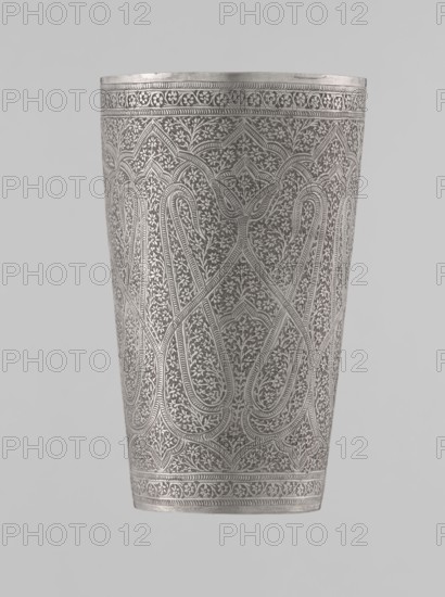 Mug, Islamic Mughal, Indian; 19th century, North India . Silver mug densely decorated with plant and floral motifs in a horror vacui style. Large symmetrically extending lines on the side wall form shapes reminiscent of flower buds. Upper and lower rims decorated with horizontal ornamental lines. Interior gilded and polished. Vessel; weight 190 g, height 12.8 cm, diameter 7.8 cm., A tall cylindrical metal vessel with intricate engraved patterns covering its surface, showing a repeating ornamental design of curved, arch-like motifs and dense foliate detailing; the vessel appears in shades of silver and gray with highlights and subtle darker gray shadows, set against a plain light gray background.