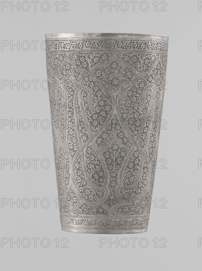 Mug, Islamic art, Mughal, Indian, horror vacui, floral ornament, plant tendril, bota ornament. Silver mug densely decorated with plant and floral motifs. Large, symmetrically extending lines on the side wall form shapes reminiscent of flower buds. The upper and lower rims are decorated with horizontal ornamental lines. The interior is gilded and polished. 19th century, North India probable, vessel; weight 190 g, height 12.8 cm, diameter 7.8 cm., A tall, cylindrical metal vessel with densely engraved, repeating floral and curving paisley-like patterns covering its surface, showing a narrow decorative band near the top and bottom; the metal has a silver-gray sheen with variations in tone where the engravings catch light, and the background is a plain light gray.