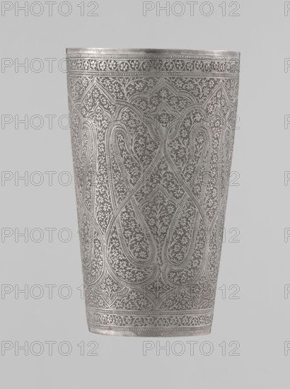 Mug, Mughal period, 19th century, North India probable. Silver mug densely decorated with plant and floral motifs in a horror vacui style. Large symmetrically extending lines on the side wall form shapes reminiscent of flower buds; upper and lower rims decorated with horizontal ornamental lines. Interior gilded and polished. Vessel, weight 190 g, height 12.8 cm, diameter 7.8 cm., A tall cylindrical metal vessel shown against a plain background with a detailed all-over engraved pattern of repeating floral and curvilinear motifs; the surface displays intricate interlacing designs with scalloped arch shapes and dense vine-like ornamentation, and there are decorative bands near the top and bottom edges; visible colors include silver, gray, and lighter highlights where the metal reflects light.