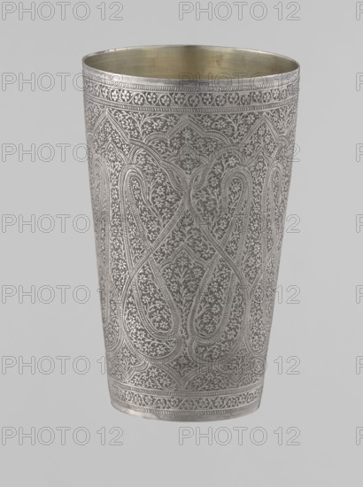 Mug, Islamic Mughal, Indian; 19th century, North India probable. Silver mug densely decorated with plant and floral motifs in a horror vacui style. Large symmetrical lines on the side wall form shapes reminiscent of flower buds; upper and lower rims decorated with horizontal ornamental lines. Interior gilded and polished. Vessel; weight 190 g; height 12.8 cm; diameter 7.8 cm., A tall metal cup with intricate fine engraving covering its exterior, showing repeating curving and floral motifs and bordered by decorative bands at the top and bottom; the exterior surface appears silver-gray while the interior shows a brass-toned golden color, and the background is a pale light gray.