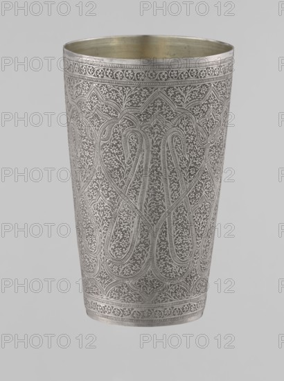 Mug, Islamic art, Mughal, Indian; 19th century, North India, silver mug densely decorated with plant and floral motifs in a horror vacui style. Large symmetrically extending lines on the side wall form shapes reminiscent of flower buds; the upper and lower rims are decorated with horizontal ornamental lines. Interior gilded and polished. Weight 190 g, height 12.8 cm, diameter 7.8 cm., A tall cylindrical metal cup with intricate raised floral and geometric patterns covering its exterior, showing repeated looping arch motifs and dense vine-like ornamentation; the rim and base have decorative bands with small floral motifs and rope-like borders. The interior appears smooth and slightly darker than the exterior. The visible colors are silver tones on the exterior and a muted goldish hue inside, set against a pale gray background.