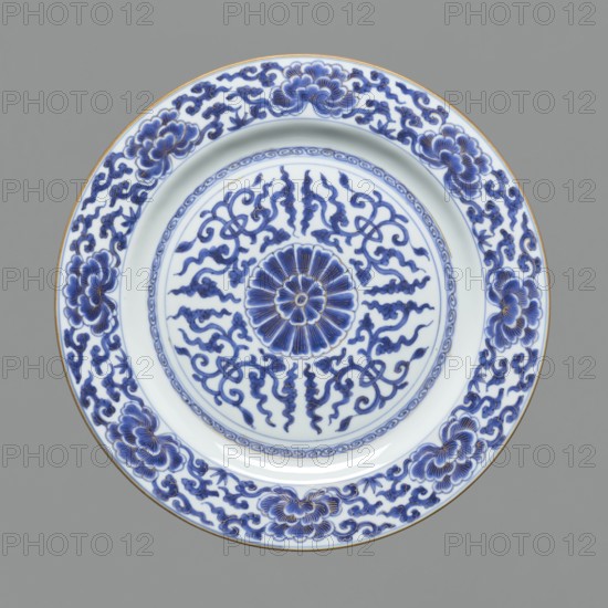 Platter, gift, Chinese art, Chinese culture, Qing 1644–1911, ceramics, blue-and-white porcelain, rosettes, floral ornaments; in the mirror in the center a stylized floral rosette. 18th century, China; dish; height 3.8 cm, diameter 34.3 cm, weight 1.35 kg, A round ceramic plate with a white base and intricate blue painted designs, centered by a floral rosette motif surrounded by swirling, ribbon-like patterns, encircled by a band of repeating decorative elements and a wide rim decorated with stylized flower clusters and scrolling vine patterns; the plate shows accents of a warm metallic color along the outer edge and the blue ranges from deep cobalt to lighter blue highlights against the white, set on a neutral gray background.