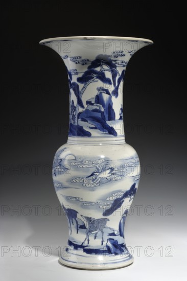 Zun-type baluster vase, porcelain, blue and white porcelain, vase with a baluster body and a long cylindrical neck flaring into a goblet shape; Kangxi style, Qing 1644–1911, Kangxi 1662–1722, dated 1662–1722; China; height 45.7 cm, diameter 23.3 cm. Decoration: deer, cranes, pines, rocks., A tall porcelain vase with a flared rim and a bulbous midsection, primarily white with detailed blue painted scenes; the upper section shows blue tree shapes and rock formations against the white background, the middle and lower sections display stylized cloud-like swirls and flying bird figures in blue, and a group of animal figures and more rocky shapes near the base; the vase sits on a flat surface with a dark background that transitions to a lighter gray near the bottom, and the visible colors are white, blue, dark gray, and light gray.