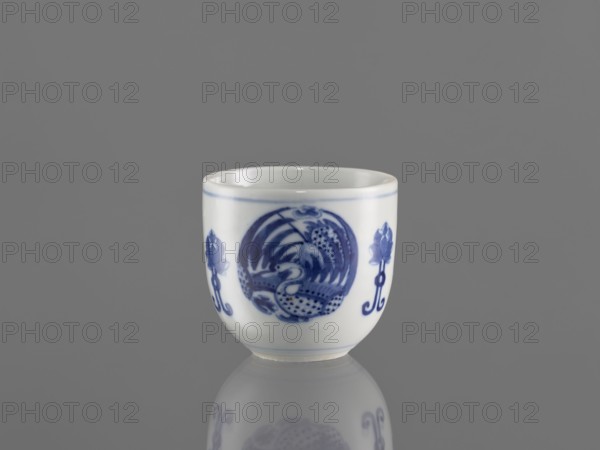 Bowl, ceramics, decorated with three phoenix images and stylized lotus flowers; blue-and-white porcelain; Chenghua style 1465–1487; Qing dynasty style 1644–1911, early 20th century Qing dynasty, 1644–1912; Chinese; vessel; height 7.5 cm, diameter 8 cm., A small white porcelain cup with a glossy finish sits against a gray background, showing a blue painted circular motif on its front flanked by blue floral-like designs; the cup has a thin blue rim near the top and a soft reflection of the cup appears on the reflective surface below.