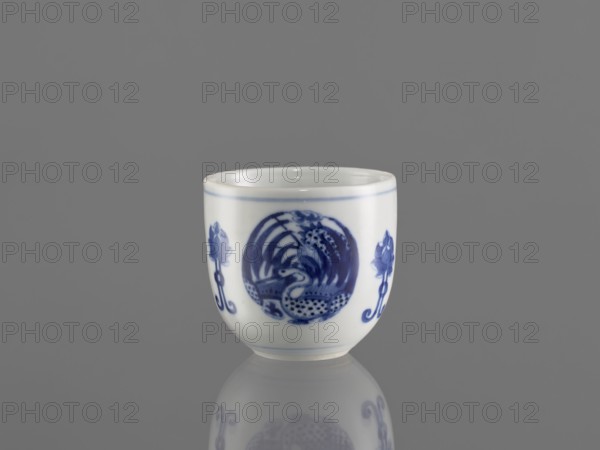 Bowl, ceramics, decorated with three phoenix images and stylized lotus flowers, blue-and-white porcelain, Chenghua period style 1465–1487, Qing dynasty style 1644–1911, early 20th century, China; vessel; height 7.5 cm, diameter 8 cm., A small white ceramic cup with blue painted decorations sits on a reflective gray surface against a gray background, showing a rounded shape and a thin blue line near the rim; the painted decoration includes a central circular blue motif with intricate patterns and flanked by stylized floral or plant motifs in blue, and the cup’s reflection appears faintly below it.