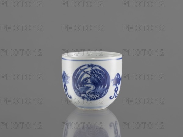 Bowl, ceramics, Chenghua 1465–1487, phoenixes and stylized lotus flowers decoration, blue-and-white porcelain, Qing style, Chinese, floral ornaments, Bowl decorated with three phoenix images and stylized lotus flowers, early 20th century Qing dynasty, 1644–1912, China, vessel, height 7.5 cm, diameter 8 cm, A small white ceramic cup with blue painted decoration sits against a gray background, showing a circular blue motif on its front flanked by floral-like blue designs on either side; the cup has a thin blue line near the rim and a subtle reflection of the cup appears on the glossy surface below.