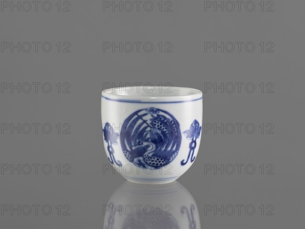 Bowl, ceramics, Chenghua 1465–1487, phoenixes and lotus iconography, blue-and-white porcelain in Qing style, floral ornaments, Bowl decorated with three phoenix images and stylized lotus flowers, early 20th century Qing dynasty, 1644–1912, China, vessel, height 7.5 cm, diameter 8 cm., A small white ceramic cup with blue painted decorative motifs sits centered on a reflective gray surface against a gray background; the cup shows a band of blue near the rim and a blue circular design on its front flanked by floral-like blue elements, and the surface reflection beneath the cup mirrors its shape and blue patterns.