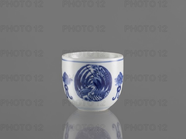 Bowl, ceramics, Chenghua 1465–1487; decorated with three phoenixes and stylized lotus flowers, blue-and-white porcelain in Qing style; early 20th century Qing dynasty, 1644–1912, China; height 7.5 cm, diameter 8 cm., A small white ceramic cup with blue painted decoration sits against a gray background and is reflected faintly beneath it; the cup has a thin blue rim and a central circular blue motif flanked by stylized floral elements, all rendered in shades of blue on the white surface.